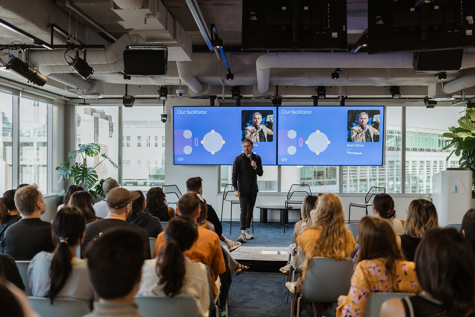 Grant Show stands on stage facilitating a design talk in front of a seated audience, with two large screens behind introducing him as the facilitator.