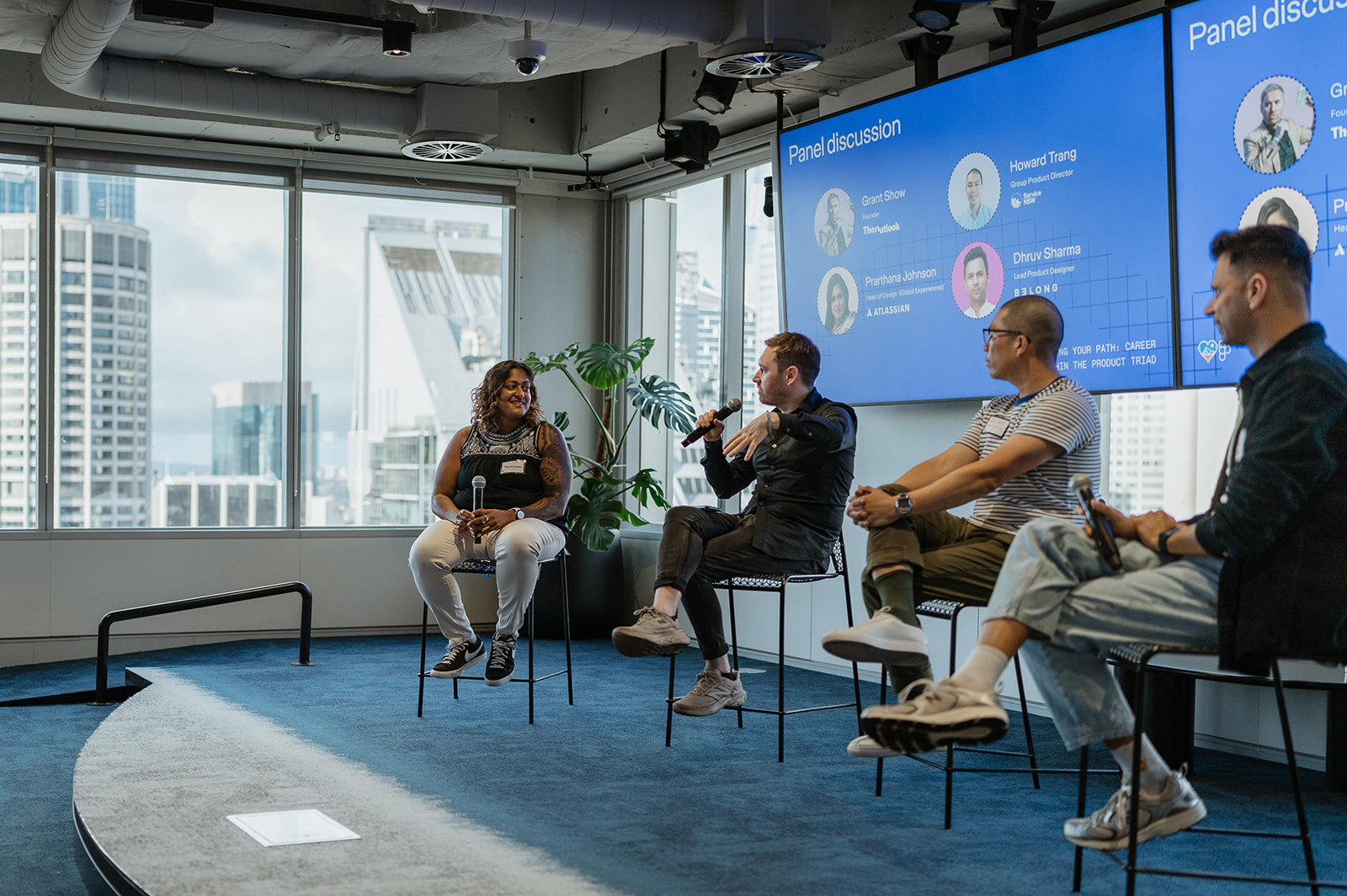 Grant Show moderates a panel discussion with four speakers seated on stage, engaging in conversation beneath a screen displaying their names and roles.